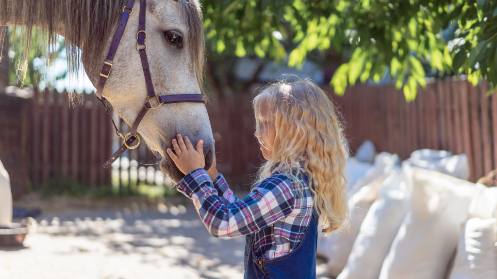 Child wearing a blue and red check shirt with long blonde wavy hair. She is patting a white pony in the face. Alternative therapies have great benefits 