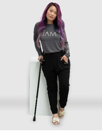 Women with purple hair with a grey top and black pants holding a cane. 