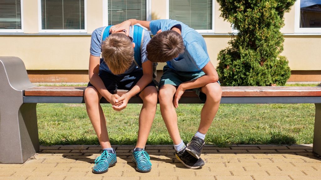 Two school kids on a bench. They have their heads in their laps.